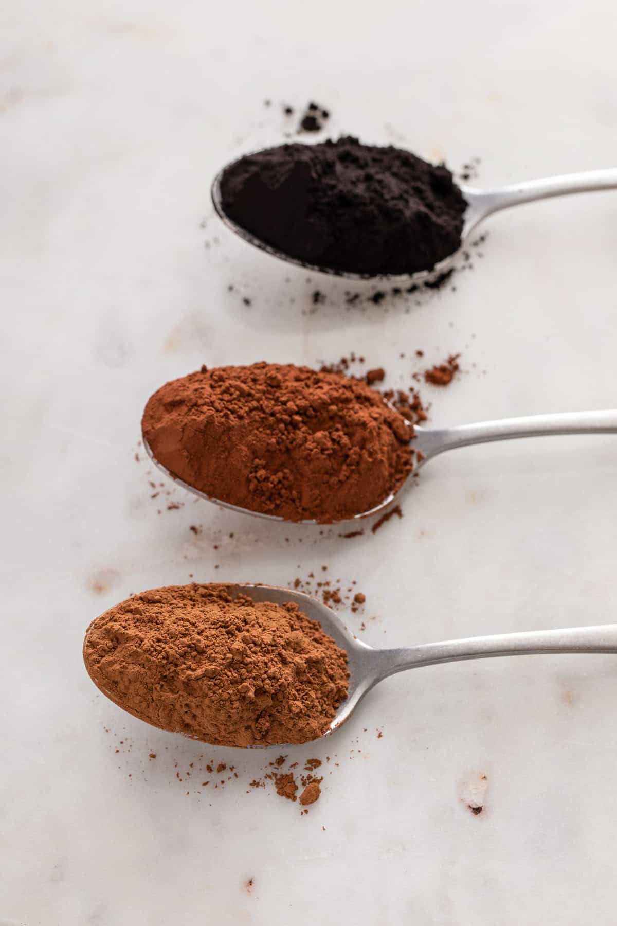 Three spoons, each holding a spoonful of cocoa powder. From the bottom up, they are: natural cocoa, dutched cocoa, and black cocoa.