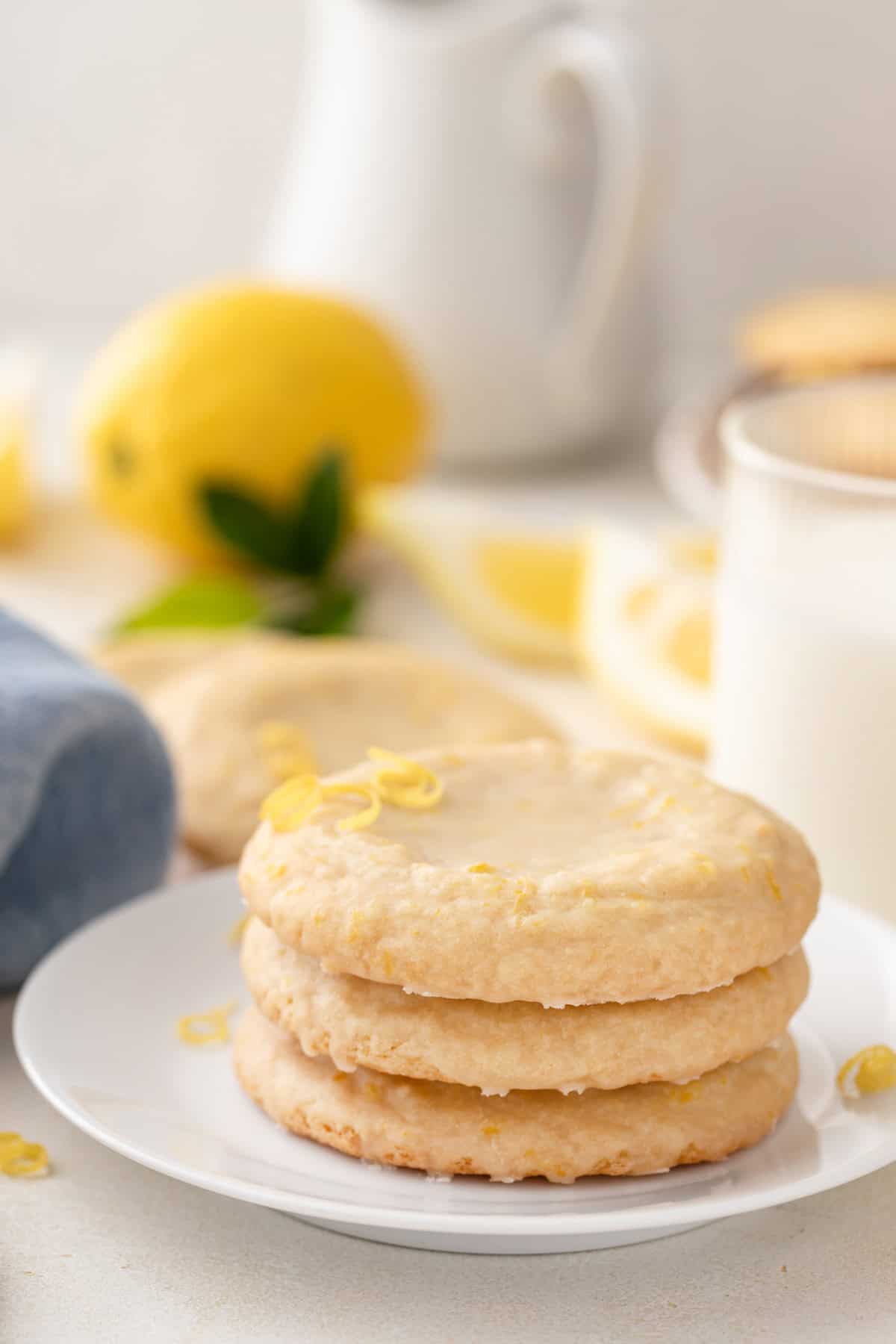 Three glazed lemon cookies stacked on a white plate in front of a glass of milk.