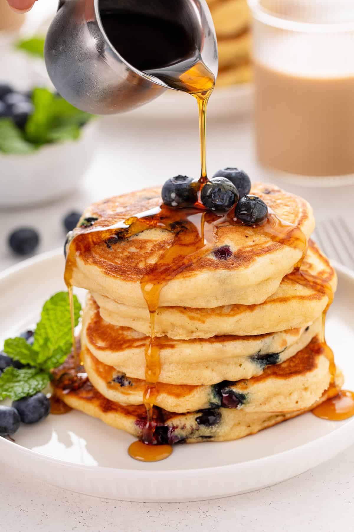 Syrup being poured over a stack of blueberry pancakes on a white plate.