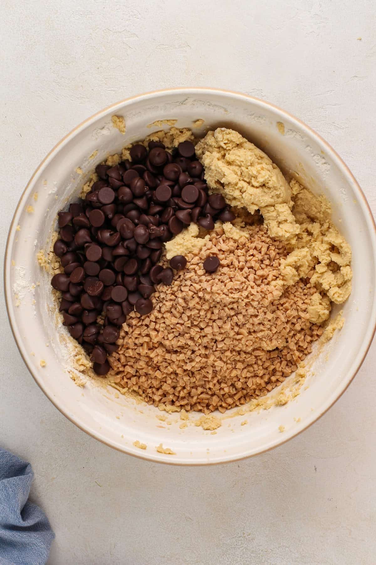 Toffee bits and chocolate chips being added to cookie dough in a mixing bowl.