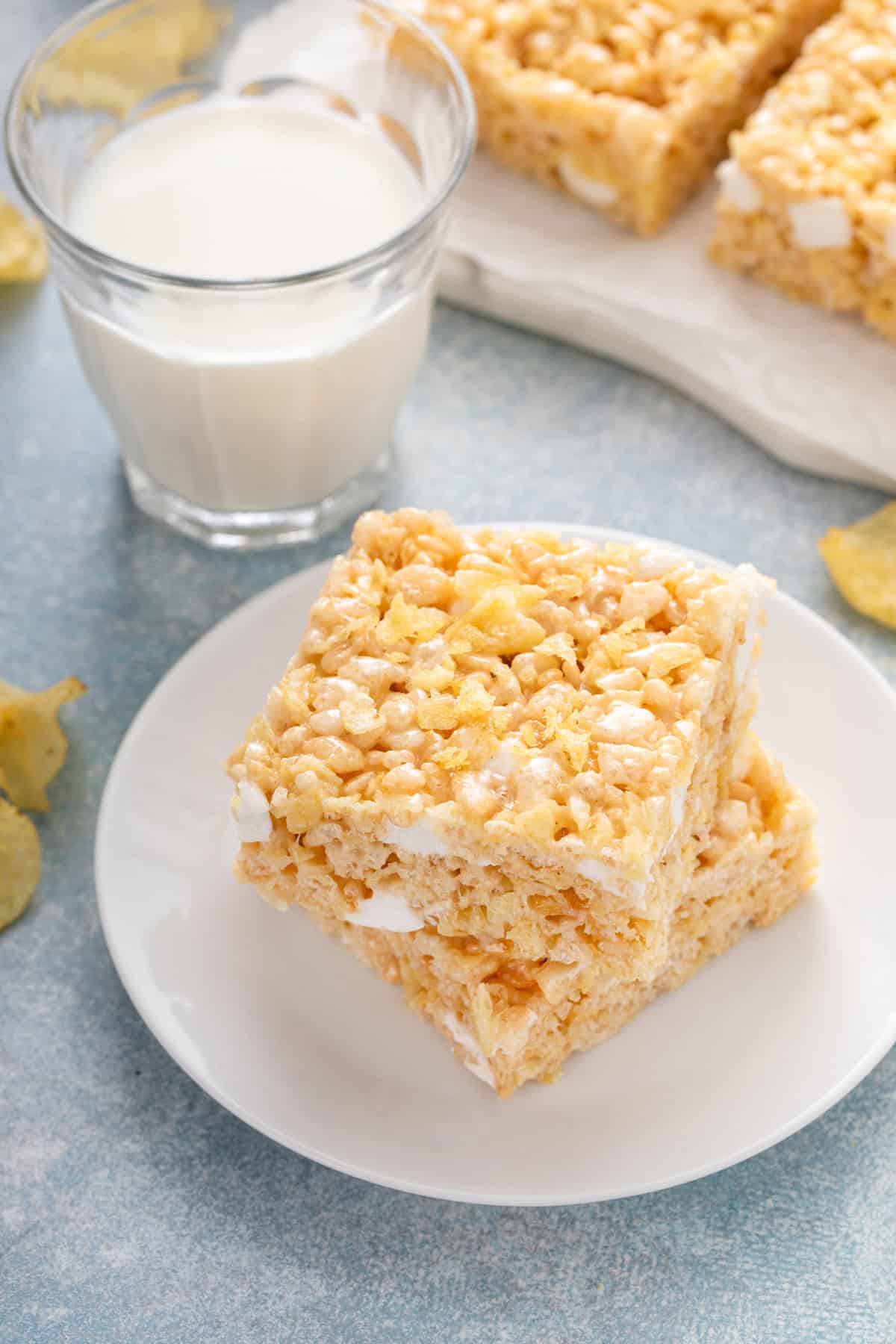 Two potato chip rice krispie treats stacked on a white plate next to a glass of milk.