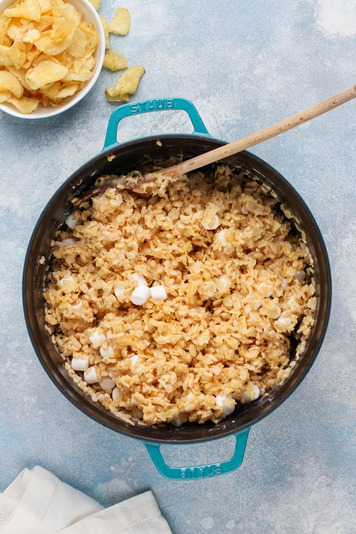 Wooden spoon stirring potato chip rice krispie treat mixture in a blue dutch oven.