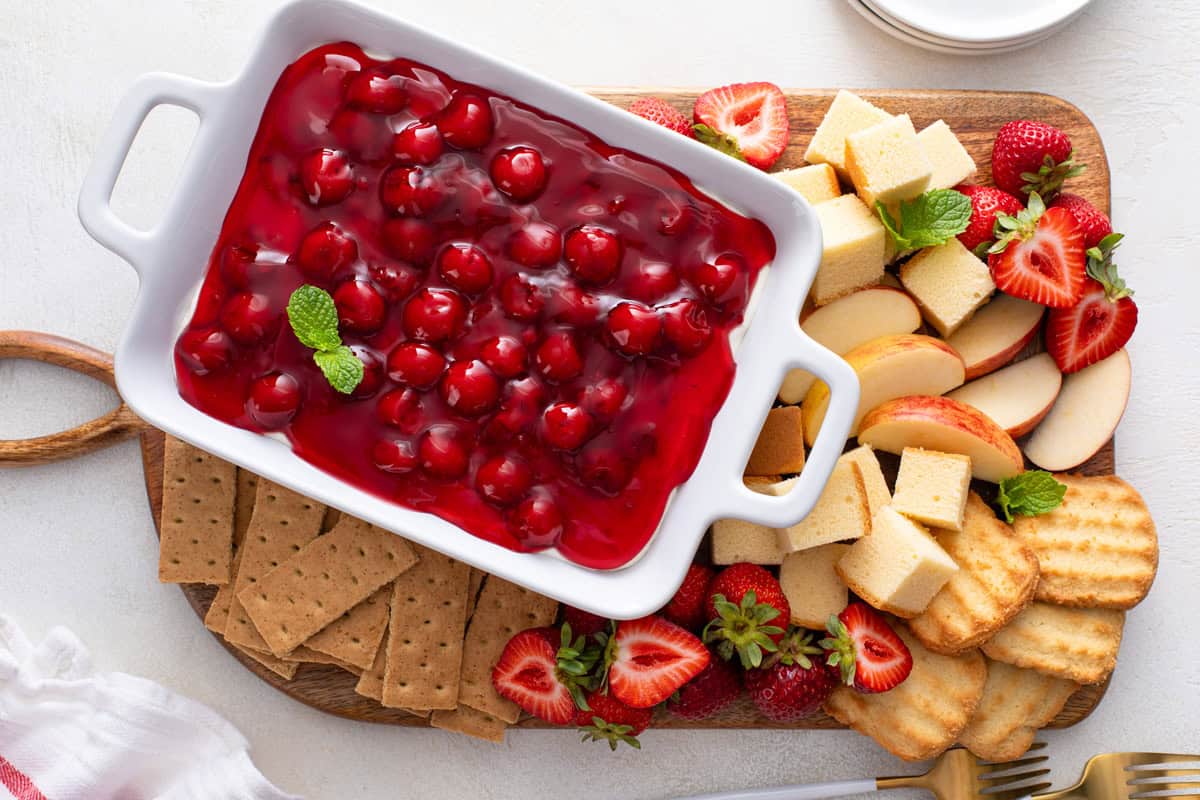 Cherry cheesecake dip in a serving dish on a wooden platter, surrounded by dippers such as cake cubes, apples, strawberries, and graham crackers.