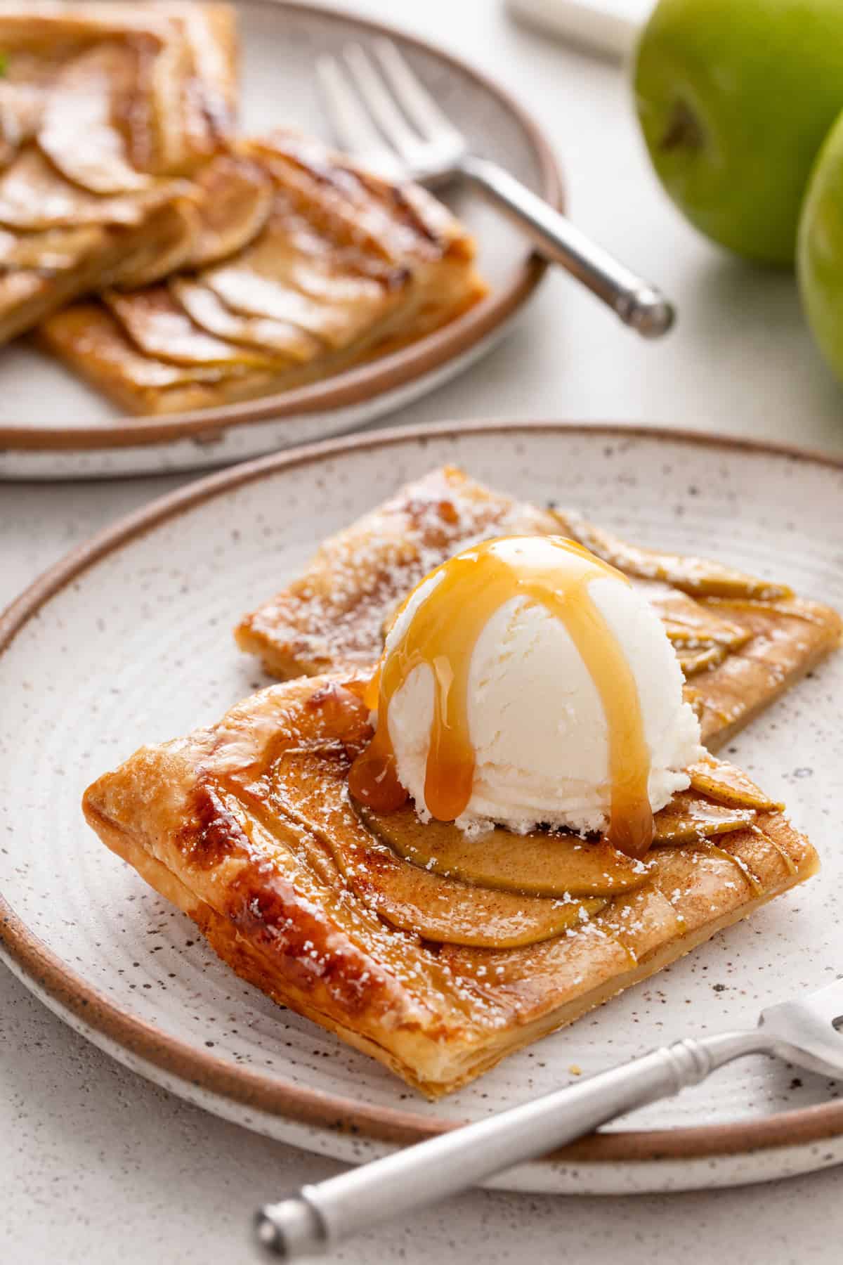 Two plated slices of puff pastry apple tart topped with a scoop of vanilla ice cream and salted caramel sauce.