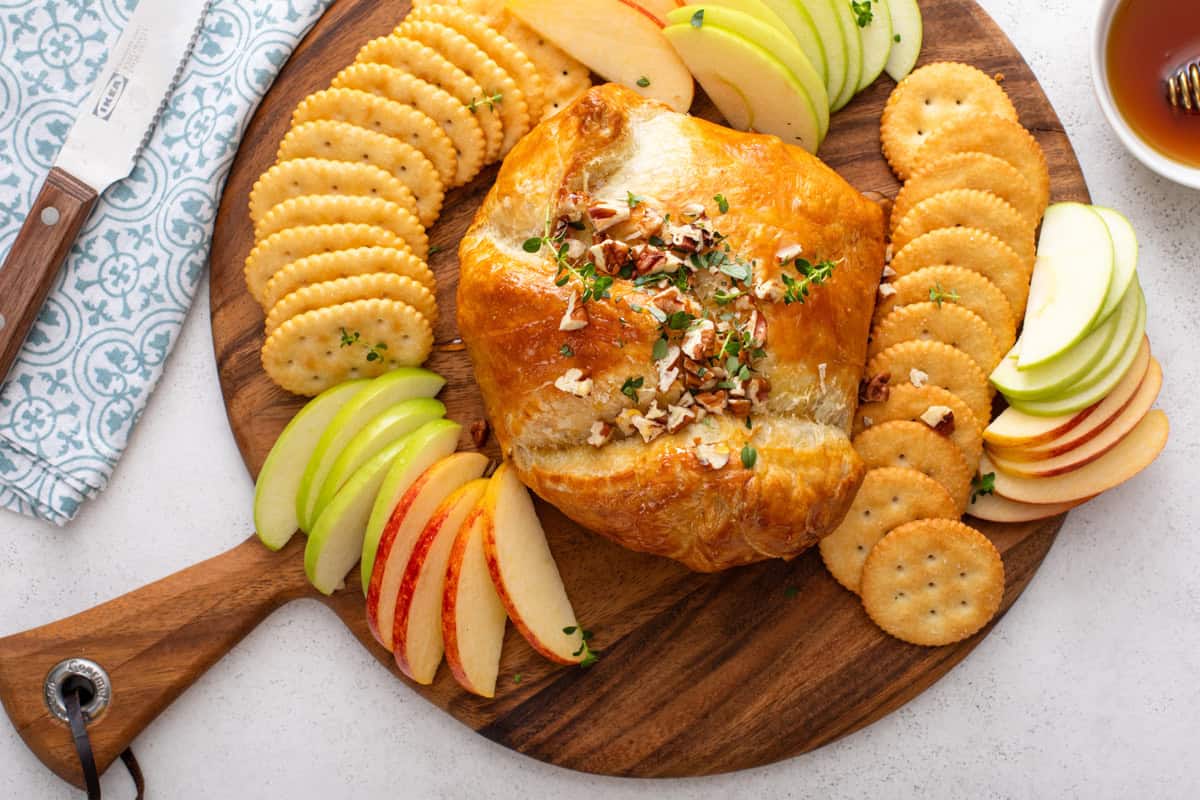 Baked brie in puff pastry on a wooden board surrounded by crackers and apples.