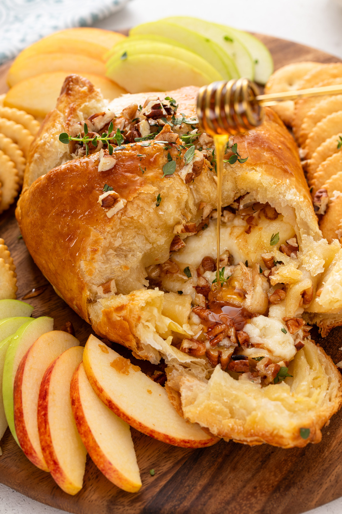 Baked brie in puff pastry that has been cut into on a board with apples and crackers. Honey is being drizzled over the cheese.