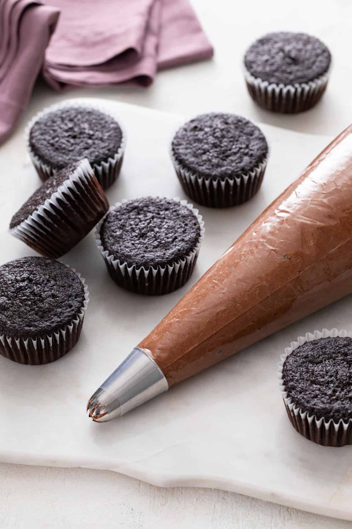 Piping bag filled with chocolate buttercream surrounded by chocolate cupcakes.