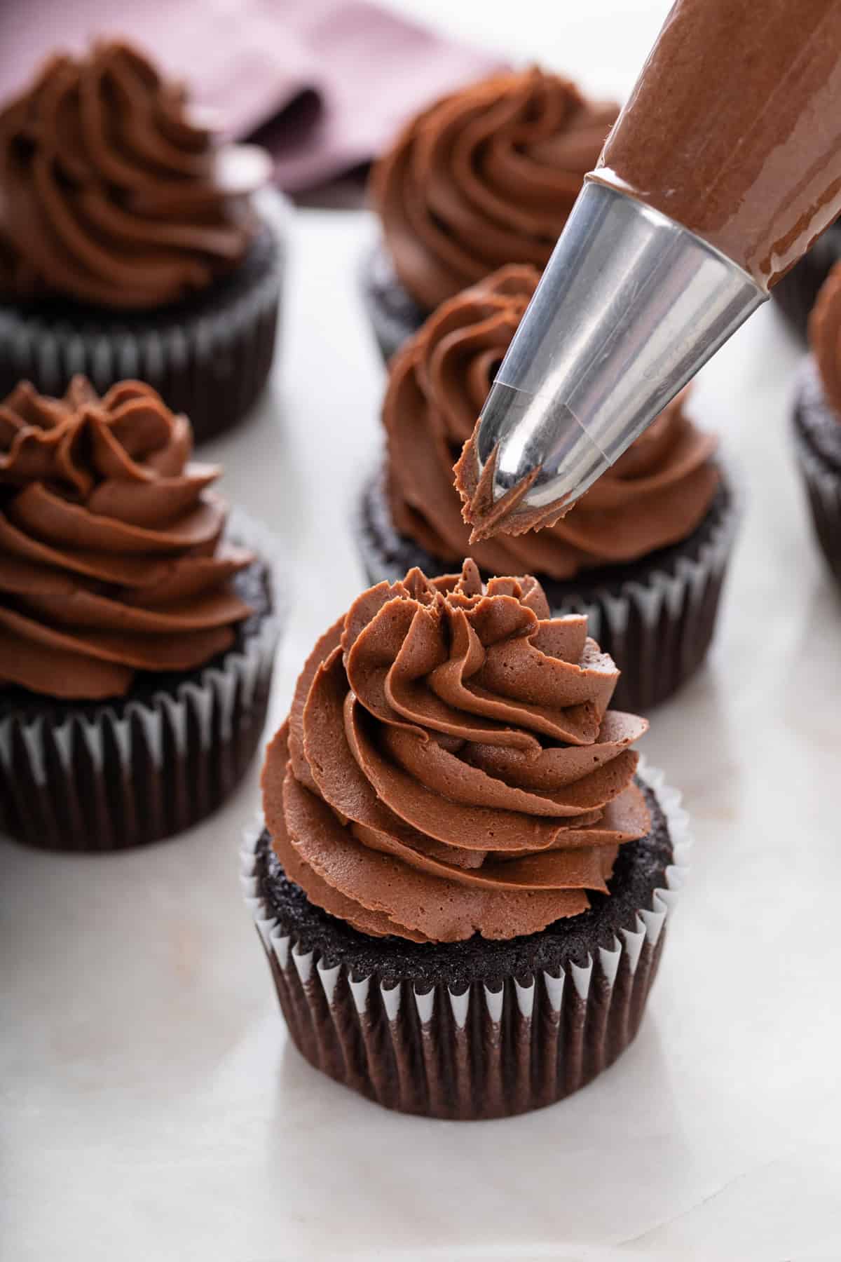 Piping bag piping chocolate buttercream on top of a chocolate cupcake.