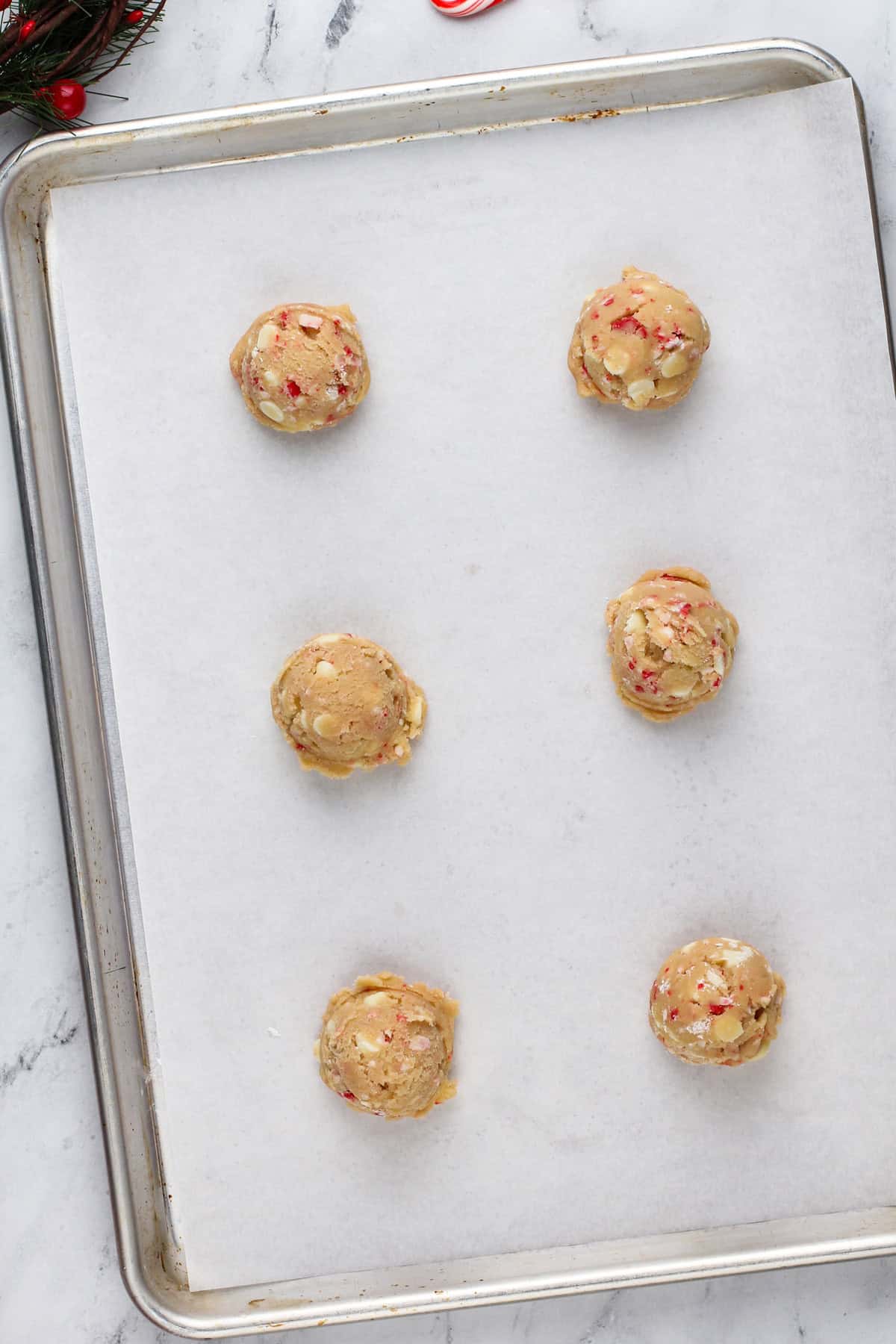 Six balls of white chocolate chip cookie dough on a lined baking sheet.