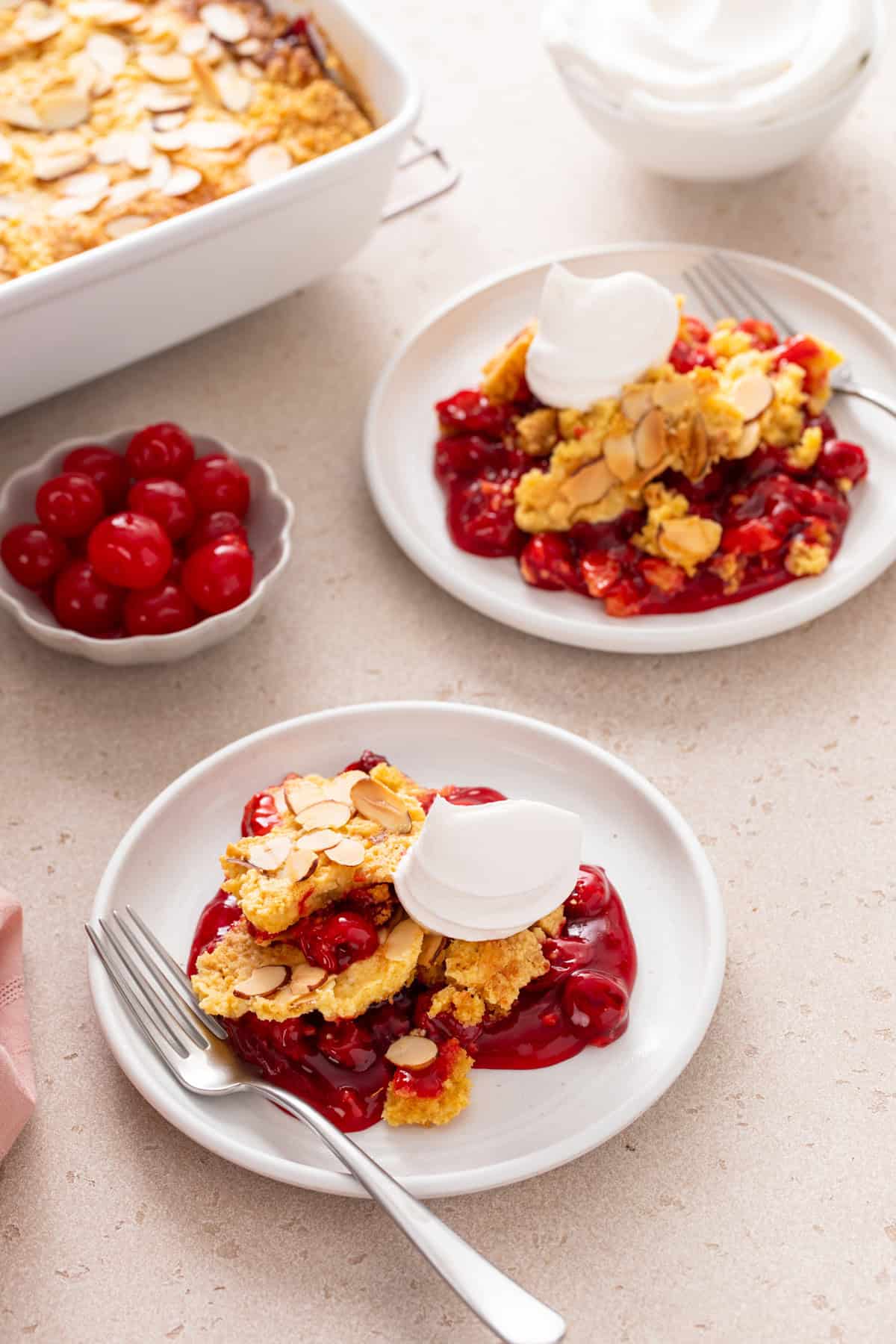 Two white plates of cherry dump cake topped with whipped cream.