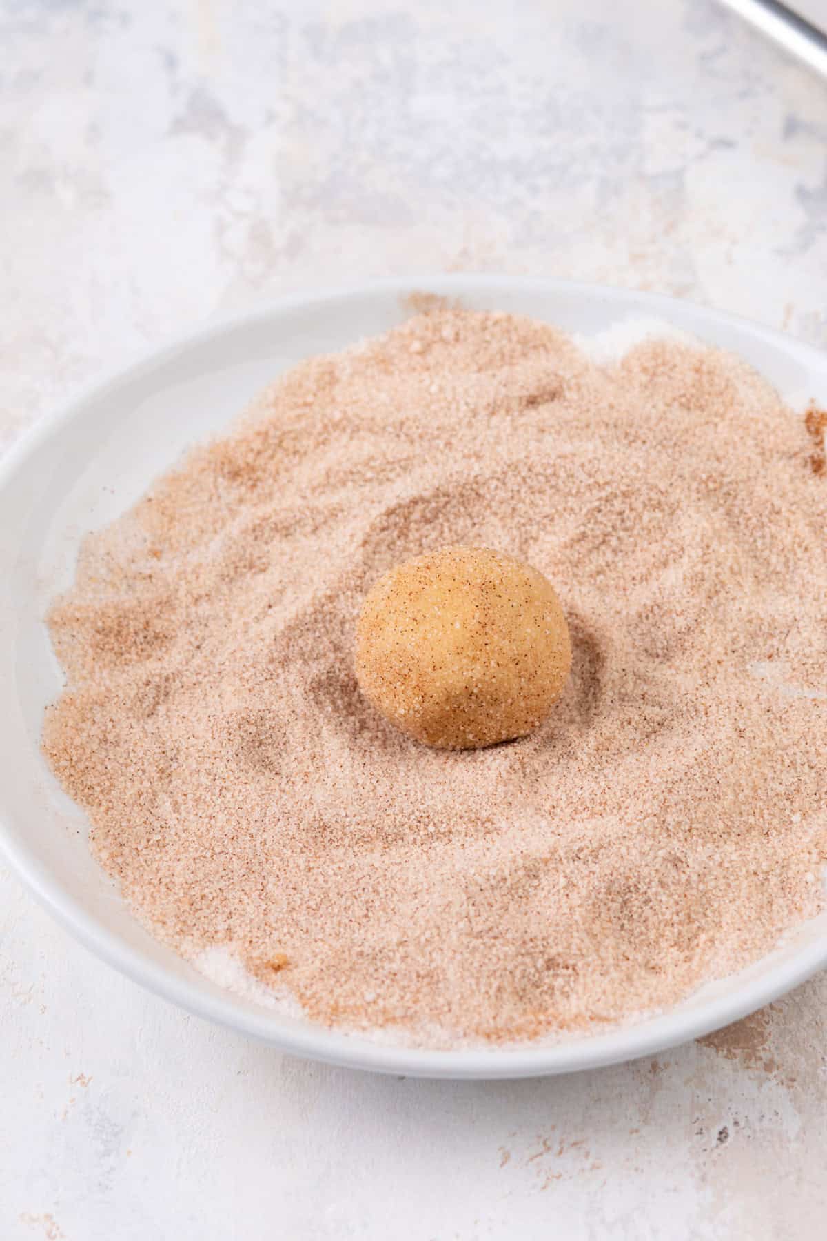 Brown butter snickerdoodle cookie dough ball being rolled in cinnamon sugar.