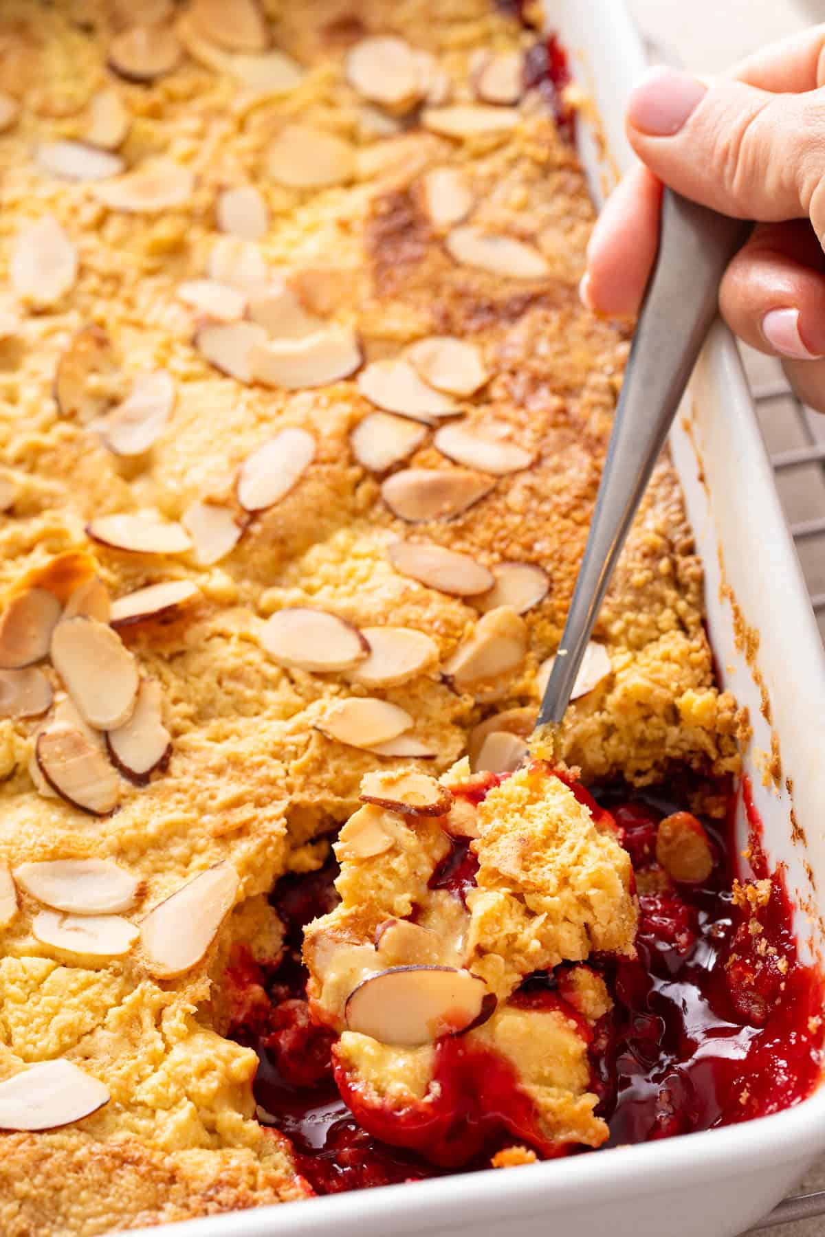 Spoon scooping cherry dump cake out of a white baking dish.