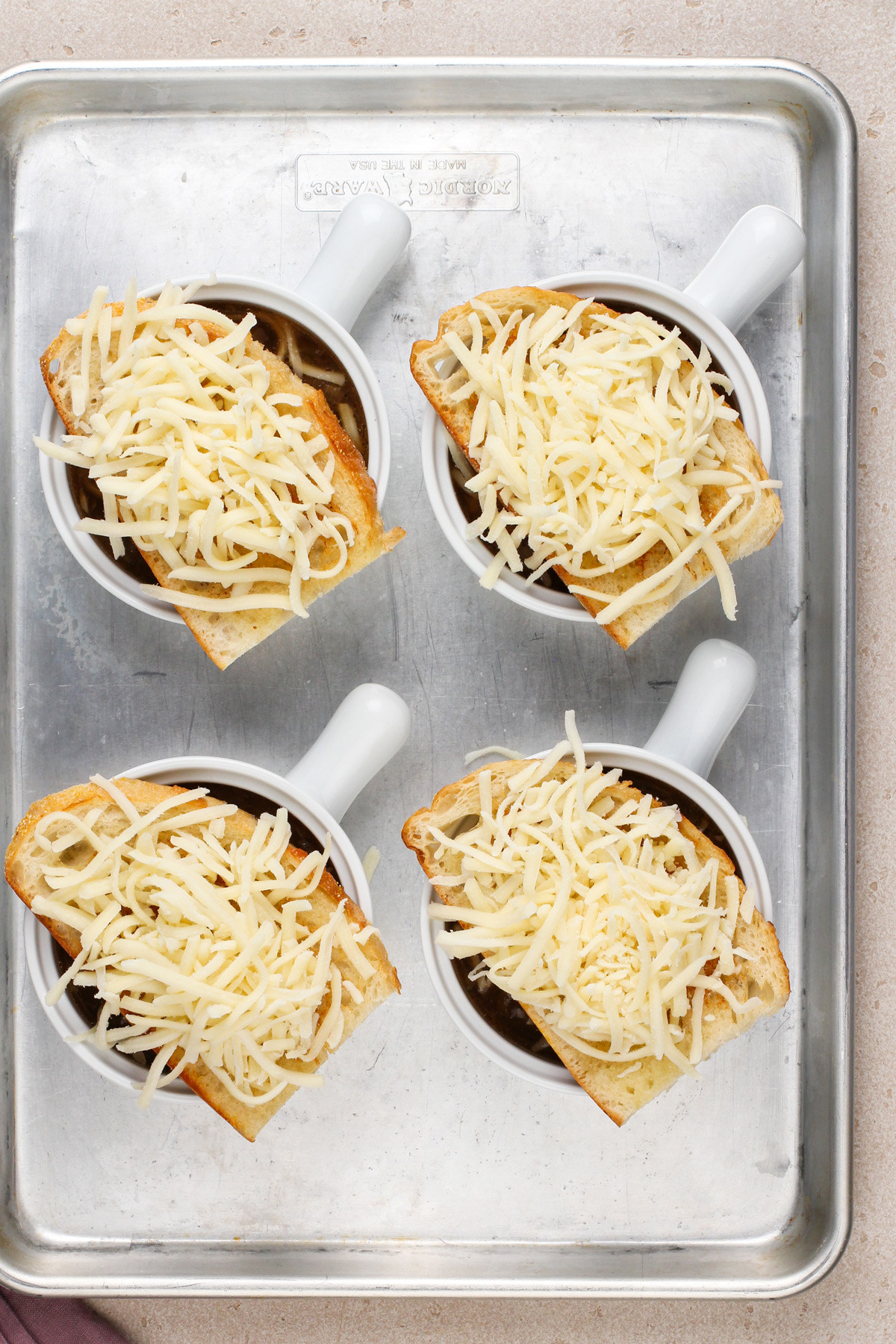 Four bowls of french onion soup assembled on a sheet tray, ready to go under the broiler.