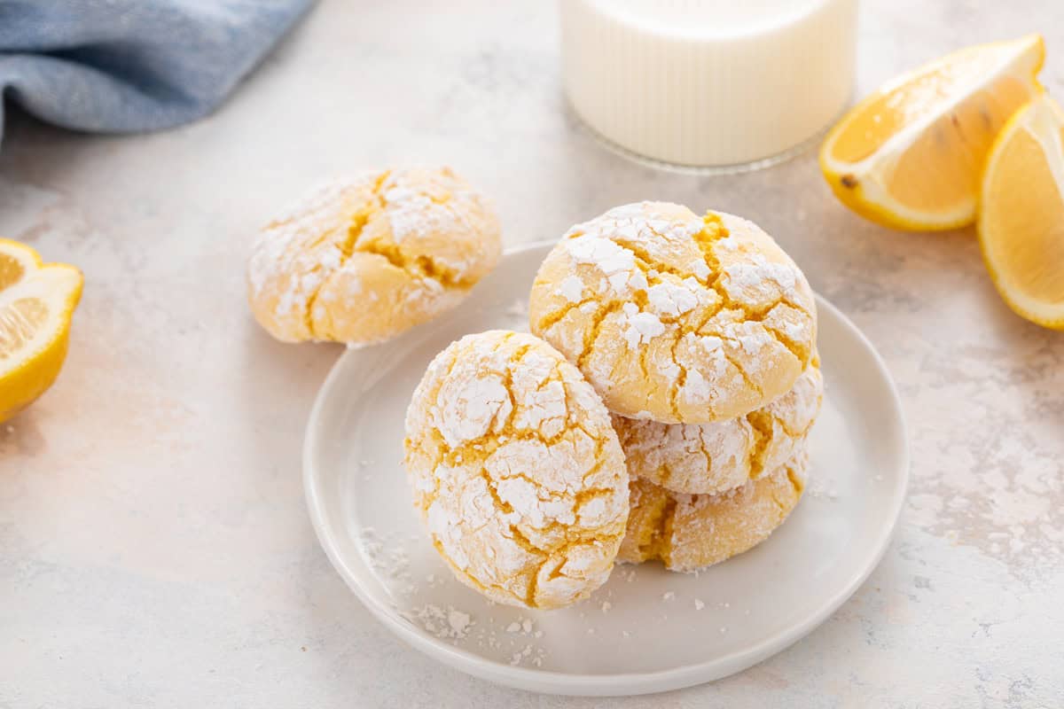 Three lemon crinkle cookies stacked on a white plate, with a fourth cookie leaning against the stack.