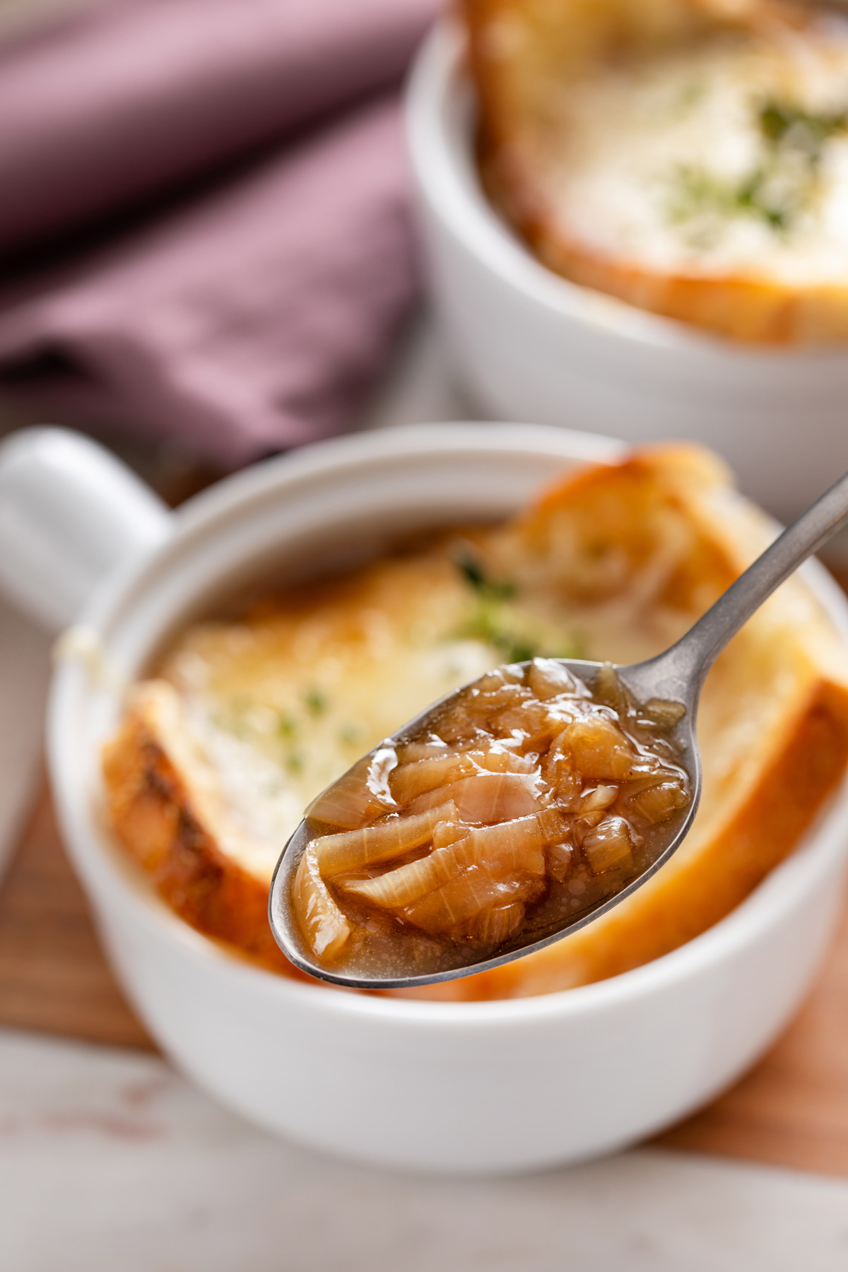 Spoon holding up a bite of french onion soup, with a bowl in the background.