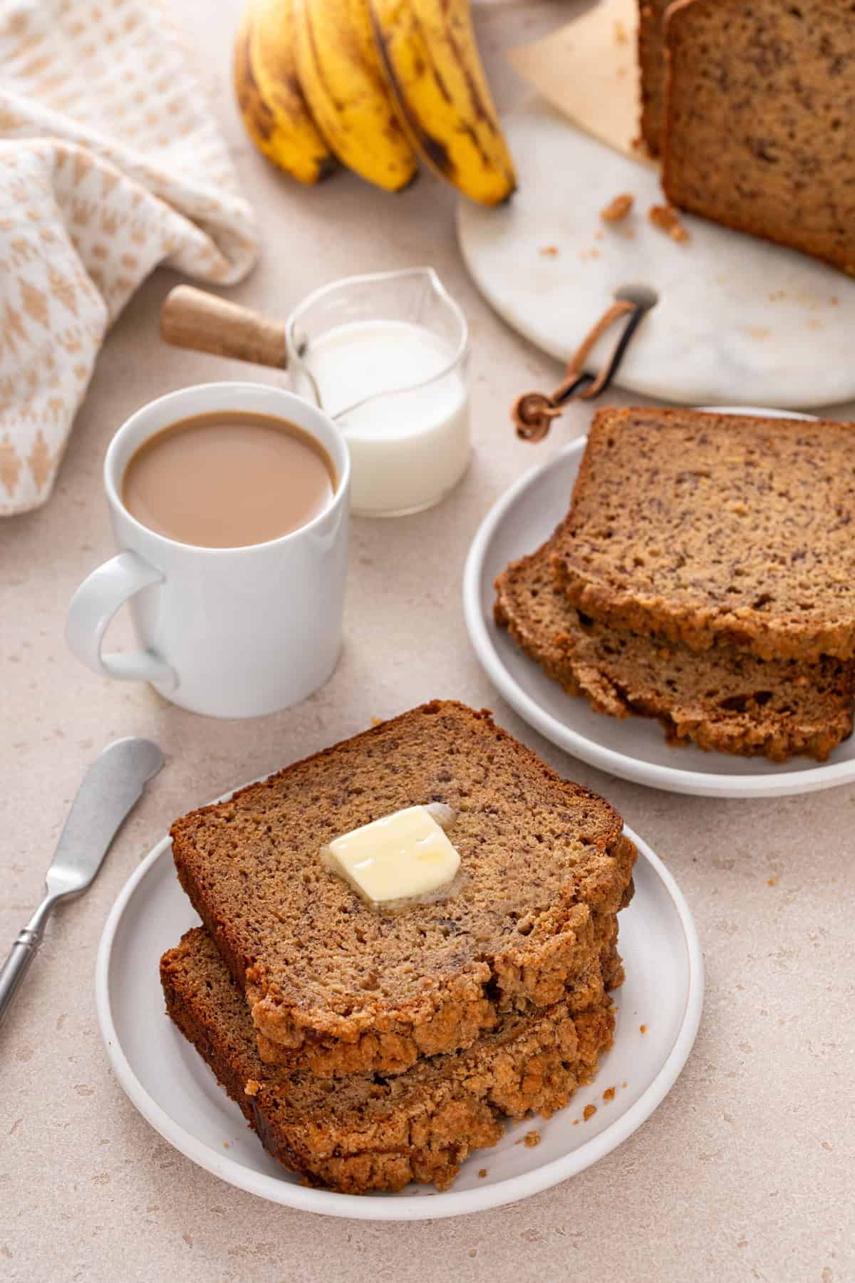 Buttered slices of sour cream banana bread on white plates next to a cup of coffee.