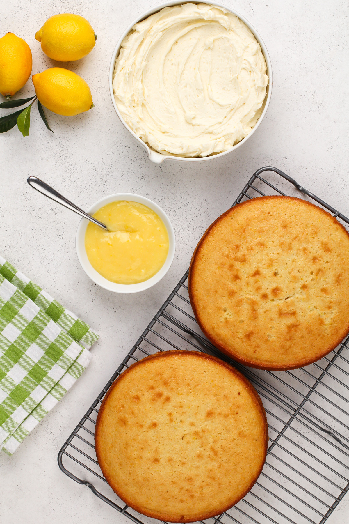 Two cooled lemon cake layers on a wire rack next to a bowl of lemon curd and a bowl of lemon frosting.