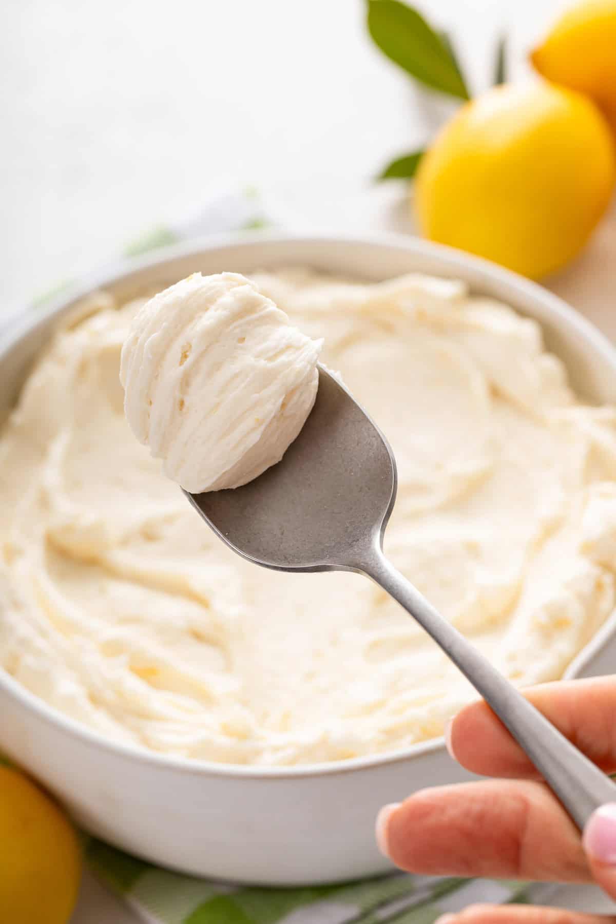 Spoon holding up a bite of lemon buttercream frosting with a bowl of the frosting in the background.